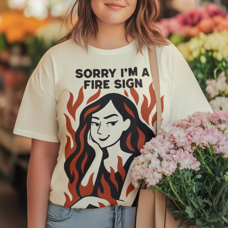 Puede incluir: Camiseta color crema con el texto "SORRY I'M A FIRE SIGN" sobre un gr&aacute;fico de una mujer con cabello negro y llamas rojas. La camiseta es de estilo casual, adecuada para el uso diario.