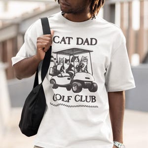May include: White t-shirt with a black and white graphic of cats riding in a golf cart. The text "Cat Dad Golf Club" is printed below the graphic.