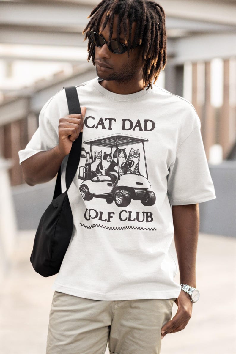 May include: White t-shirt with a black and white graphic of cats riding in a golf cart. The text "Cat Dad Golf Club" is printed below the graphic.