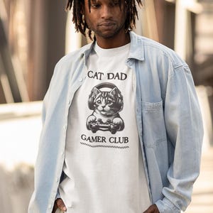 May include: White t-shirt with a graphic of a cat wearing headphones and holding a game controller. The text "CAT DAD" is above the cat, and "GAMER CLUB" is below. The shirt is worn by a person wearing a light blue button-down shirt.