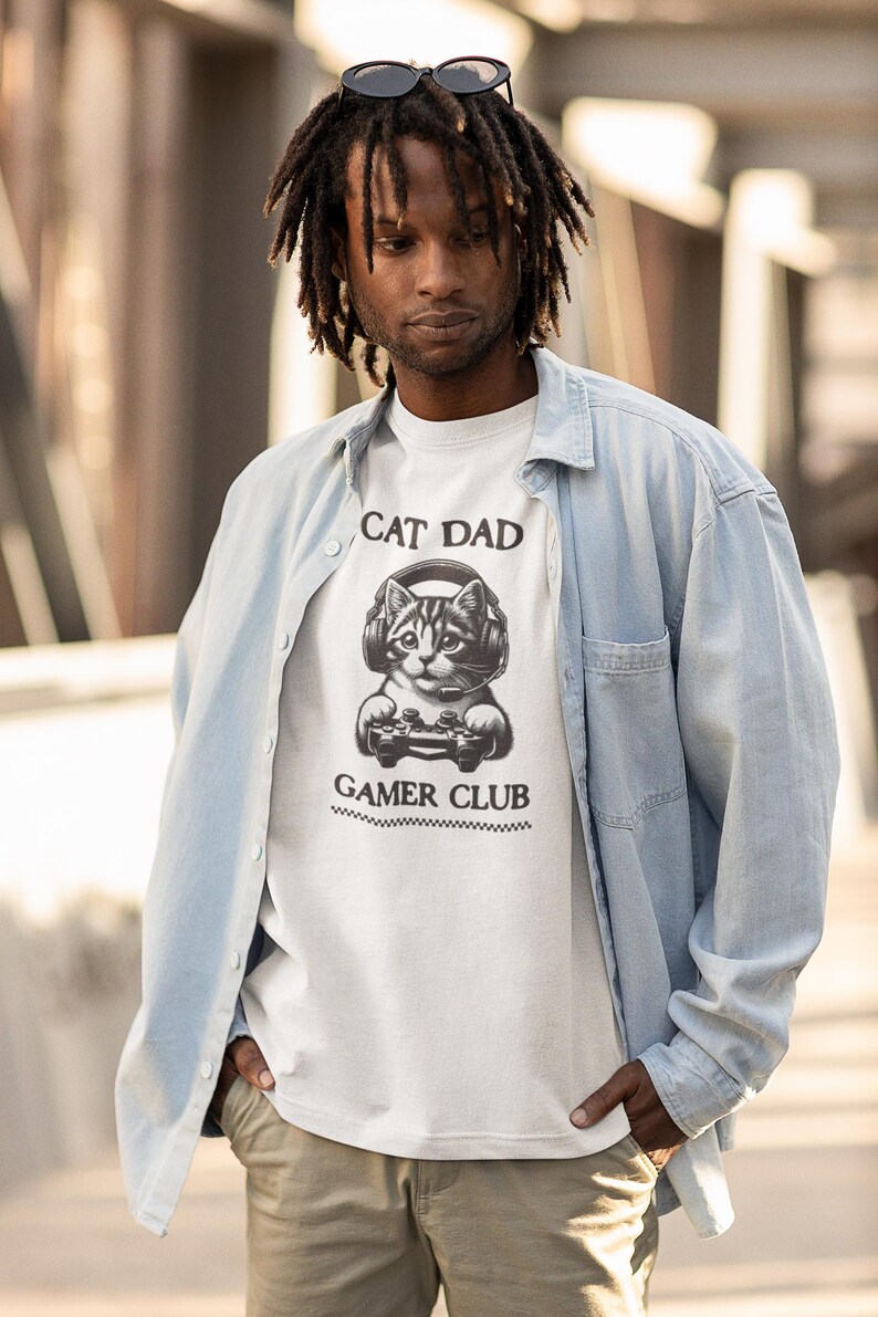 May include: White t-shirt with a graphic of a cat wearing headphones and holding a game controller. The text "CAT DAD" is above the cat, and "GAMER CLUB" is below. The shirt is worn by a person wearing a light blue button-down shirt.
