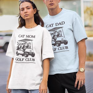 May include: A couple wearing matching white and light blue t-shirts with a graphic of cats riding in a golf cart. The shirts say "Cat Mom Golf Club" and "Cat Dad Golf Club".