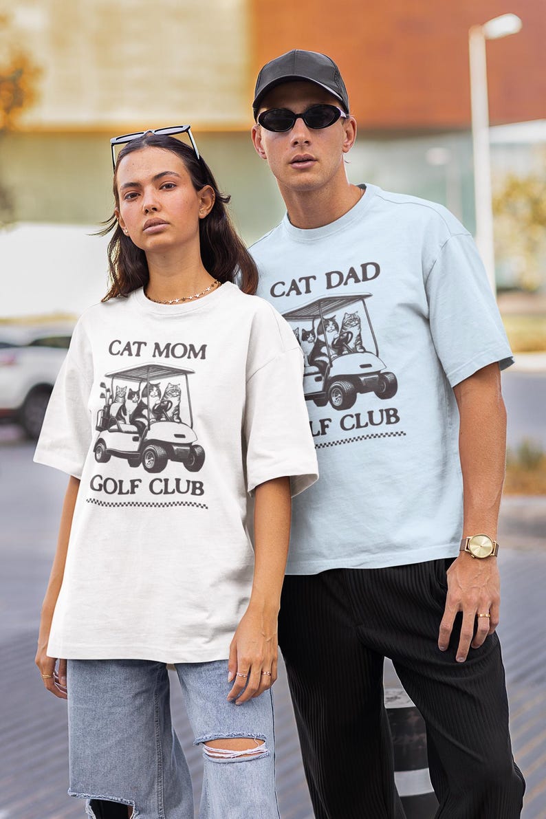 May include: A couple wearing matching white and light blue t-shirts with a graphic of cats riding in a golf cart. The shirts say "Cat Mom Golf Club" and "Cat Dad Golf Club".