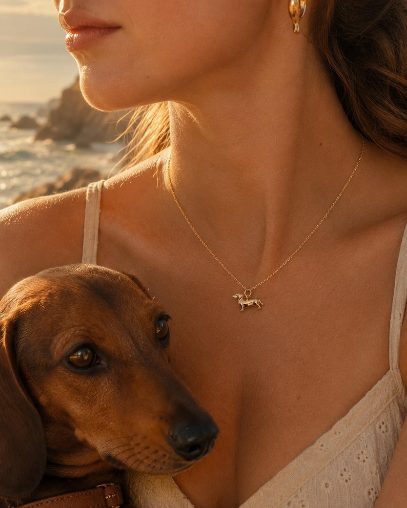 May include: A gold dachshund pendant necklace. The dog charm hangs from a delicate chain. The necklace is worn by a person wearing a cream-coloured top. A brown dachshund dog is in the foreground.