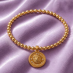 May include: Gold-colored beaded bracelet with a lion pendant. The pendant is round with a detailed lion's head and small, sparkling accents. The bracelet is set against a soft, lavender-colored fabric.