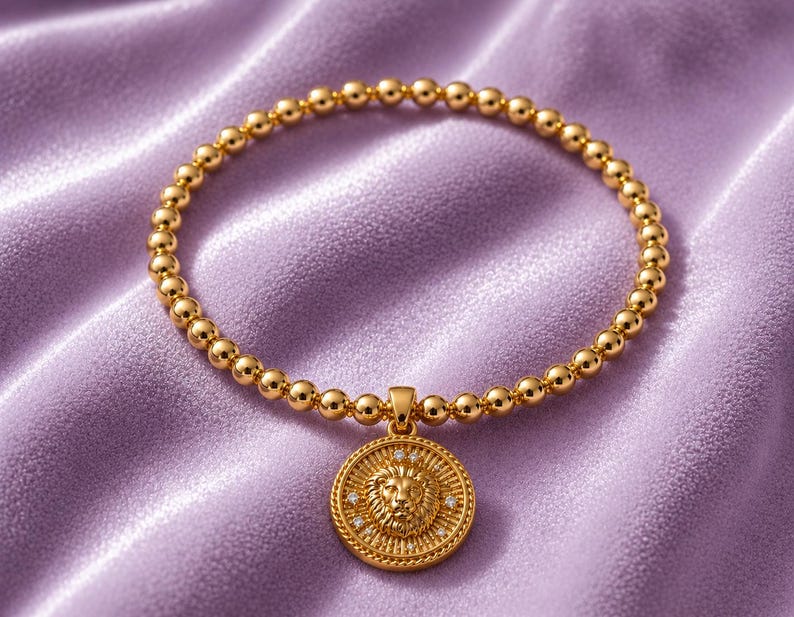 May include: Gold-colored beaded bracelet with a lion pendant. The pendant is round with a detailed lion's head and small, sparkling accents. The bracelet is set against a soft, lavender-colored fabric.