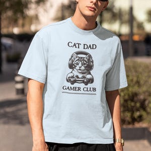 May include: Light blue t-shirt with the text "CAT DAD GAMER CLUB" and a graphic of a cat wearing headphones and holding a game controller. The shirt is a short-sleeved crew neck.