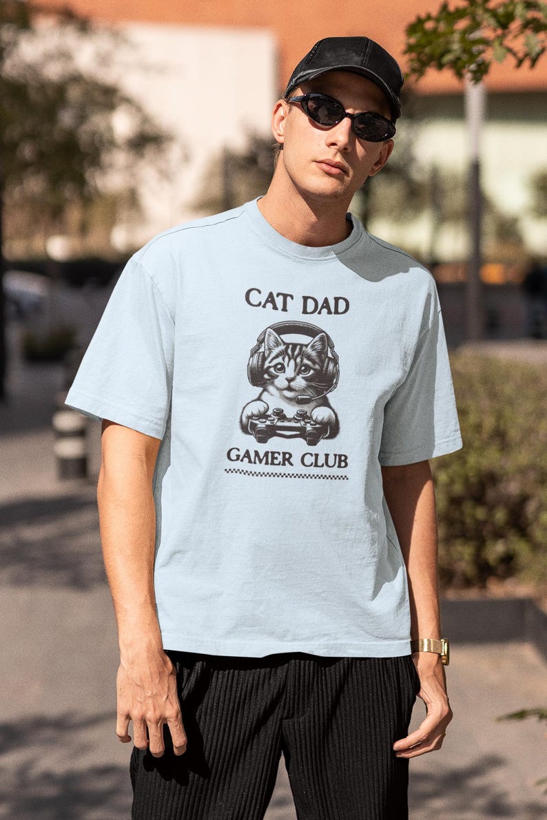 May include: Light blue t-shirt with the text "CAT DAD GAMER CLUB" and a graphic of a cat wearing headphones and holding a game controller. The shirt is a short-sleeved crew neck.