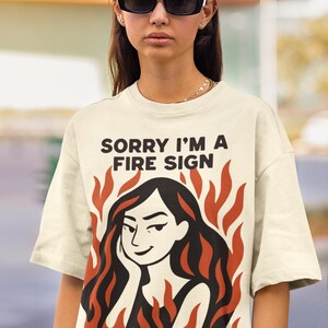 Puede incluir: Camiseta color crema con el texto negro "SORRY I'M A FIRE SIGN". La camiseta presenta un gr&aacute;fico de una mujer con pelo largo negro rodeada de llamas naranjas. La persona lleva gafas de sol negras.