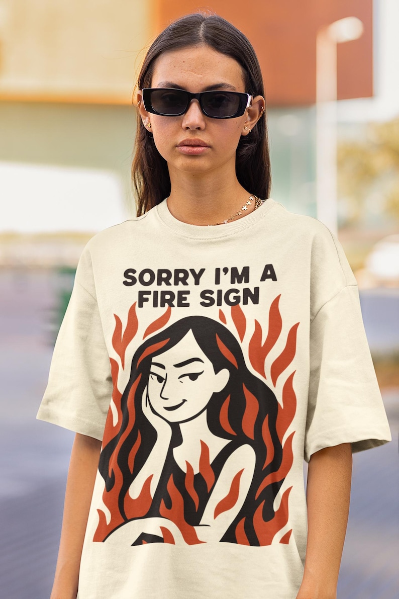 Puede incluir: Camiseta color crema con el texto negro "SORRY I'M A FIRE SIGN". La camiseta presenta un gr&aacute;fico de una mujer con pelo largo negro rodeada de llamas naranjas. La persona lleva gafas de sol negras.