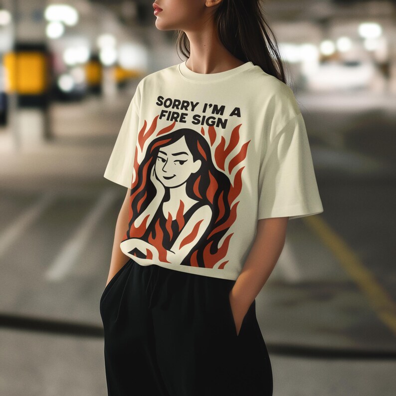 Puede incluir: Camiseta color crema con el texto "SORRY I'M A FIRE SIGN". La camiseta presenta un gr&aacute;fico de una mujer con cabello negro y llamas rojas. La modelo lleva pantalones negros.