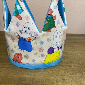 May include: A fabric crown featuring a cream-colored background with a repeating pattern of cartoon rabbits in colorful clothing, including blue overalls and yellow dresses, interspersed with small multi-colored flowers and a bright blue trim.