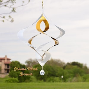 May include: A hanging wind spinner featuring a spiraling, diamond-shaped design with alternating silver and gold metallic panels. A clear, faceted crystal bead hangs from the bottom point. The text "Corona Wind Spinner" is visible on the left.
