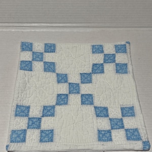 Puede incluir: Un pequeño edredón cuadrado con fondo blanco y un diseño de tablero de ajedrez azul y blanco. El edredón está hecho de tela y tiene una textura acolchada. El diseño es una serie de cuadrados dispuestos en un patrón repetitivo.