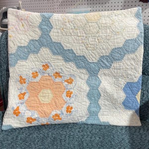 May include: A square throw pillow featuring a patchwork quilt design with a hexagonal pattern. The fabric displays a color palette of white, light blue, and soft peach, accented by small orange floral motifs stitched into the quilted surface.