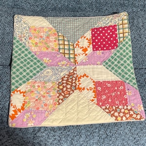 May include: A square quilted pillow cover featuring a patchwork design of various fabric patterns including floral prints, plaid, stars, and solid colors in shades of pink, orange, green, white, and brown with visible diagonal stitching throughout.