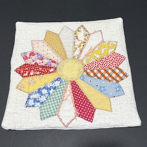 May include: A square, white quilted pillow with a large, colorful flower design. The flower's petals are made from various patterned fabrics in red, yellow, blue, and other colors. The center of the flower is a solid yellow circle. The pillow is a decorative home accent.