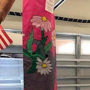 May include: A pink, yellow, white and green windsock with a watering can and flowers printed on the top section. The windsock is hanging from a golf cart.