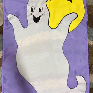 May include: A rectangular purple garden flag featuring a large white ghost with a smiling face and outstretched arms. Behind the ghost is a bright yellow full moon with a small black bat silhouette flying in front of it. The design is centered.