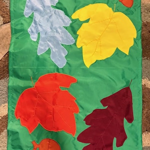May include: A rectangular green fabric garden flag featuring appliqued fabric shapes of a light blue oak leaf, a yellow maple leaf, a bright orange maple leaf, a dark maroon oak leaf, and two clusters of orange acorns stitched onto the surface.