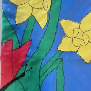 May include: A fabric art piece featuring a floral design with yellow daffodils, a red tulip, and a black butterfly against a blue and green background. The flowers and butterfly are outlined with black stitching.