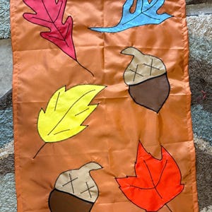 May include: A rectangular orange fabric flag featuring appliqued designs of two brown acorns with tan caps and three leaves in red, yellow, and light blue. Each element is outlined in black stitching, and the flag has a horizontal hem at the bottom.