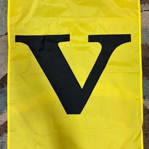 May include: A yellow flag with a large black letter "V" in the center. The flag has a yellow border and is made of a lightweight material. The flag is likely for decorative or promotional purposes.