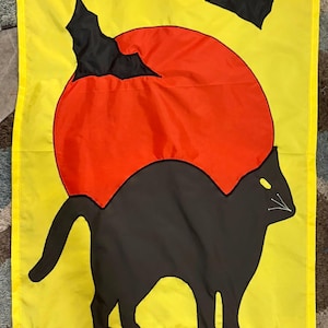 May include: A rectangular yellow garden flag featuring a large orange circular moon behind a black cat silhouette. The cat stands on a brown horizontal bar, with two black bat silhouettes flying in the upper portion of the yellow background.