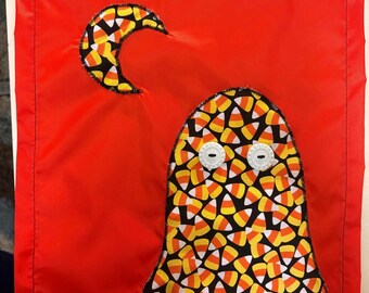 May include: A rectangular red fabric wall hanging featuring a ghost-shaped applique and a crescent moon applique. Both shapes are made from fabric with a candy corn pattern. The ghost has two white circular buttons attached as eyes.
