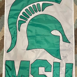 May include: A white flag featuring a green Spartan helmet logo and the letters "MSU" in green. The flag has a rectangular shape and is likely used for decorative or promotional purposes, possibly for a university or sports team.