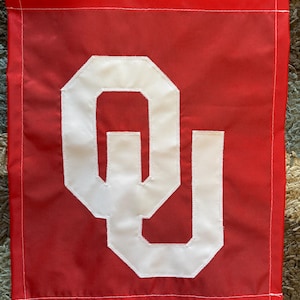 May include: A rectangular red fabric flag featuring a large, white, stylized interlocking OU logo centered on the front. The flag has white stitched borders along the top and sides, with a smooth, semi-glossy finish on the red material.