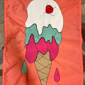 May include: A rectangular orange flag featuring a central graphic of a triple-scoop ice cream cone. The scoops are white, mint green, and hot pink, topped with a red cherry. A tan waffle cone is at the base, with small green and pink drips.