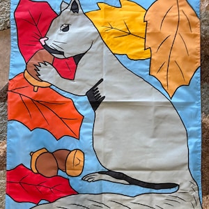 May include: A rectangular garden flag featuring a light blue background with a large gray squirrel holding a brown acorn. The design includes vibrant red, orange, and yellow autumn leaves surrounding the squirrel, with two additional brown acorns positioned near the bottom left corner.