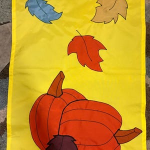 May include: A rectangular yellow fabric wall hanging featuring a decorative applique design of two orange pumpkins at the bottom, accented by a dark purple leaf, and three falling leaves in blue, tan, and red colors positioned above the pumpkins.