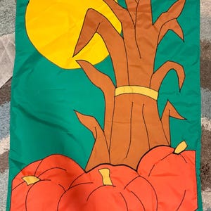 May include: A rectangular fabric garden flag featuring a green background with a large yellow sun, a brown corn stalk tied with a yellow band, and three orange pumpkins with yellow stems at the base, all outlined in black stitching.