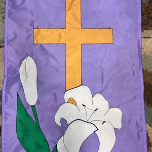 May include: A purple garden flag featuring a gold cross, white lilies, and a white tulip. The flag has a purple border and is made of a lightweight fabric. The design is a religious theme.