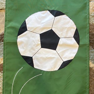 May include: A rectangular green fabric flag featuring a large, centered soccer ball with classic black and white pentagonal and hexagonal panels. Two thin, curved white lines are stitched onto the lower left section of the green fabric.