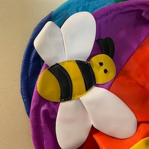 May include: A colorful, rainbow-themed decoration featuring a large, stylized bee. The bee has a yellow body with black stripes and white wings. The background is a vibrant array of colors, including blue, purple, red, and yellow.