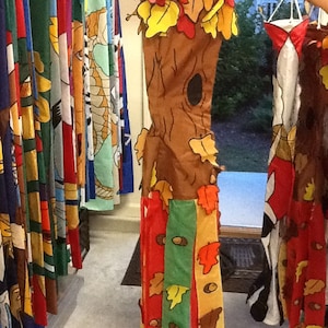 May include: A decorative fall-themed windsock shaped like a tree trunk with colorful leaves. The windsock is brown with a hollowed-out section and attached leaves in red, yellow, and orange. Other colorful windsocks hang in the background.