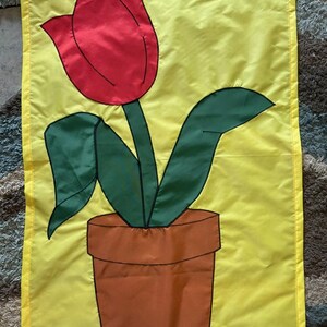 May include: A rectangular yellow garden flag featuring a central applique design of a red tulip flower with green leaves planted in a brown terracotta-colored flower pot. The design is outlined in black stitching on a smooth, solid yellow fabric background.