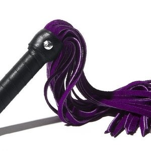 May include: A purple velvet flogger with a black handle. The flogger is made of multiple strands of velvet that are tied together at the end of the handle.