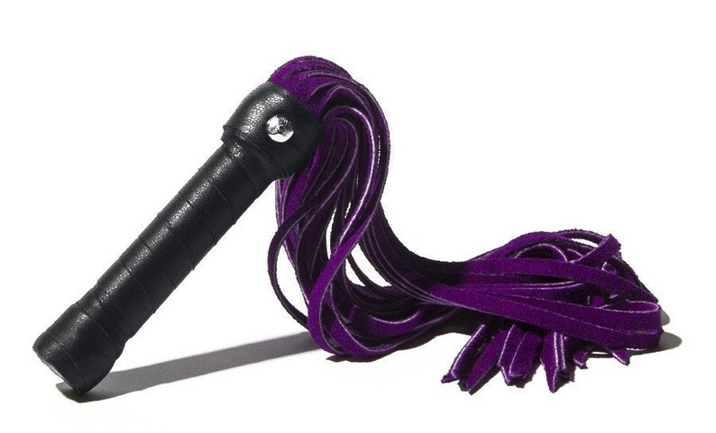 May include: A purple velvet flogger with a black handle. The flogger is made of multiple strands of velvet that are tied together at the end of the handle.