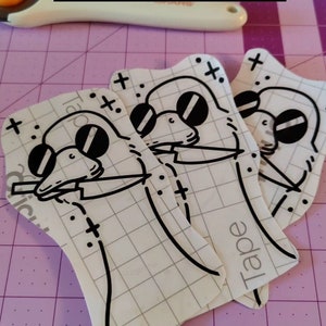 May include: Three white vinyl stickers featuring a cartoon duck wearing sunglasses and holding a knife. The stickers have a grid pattern and the word "Tape" printed on them. The stickers are on a purple cutting mat.