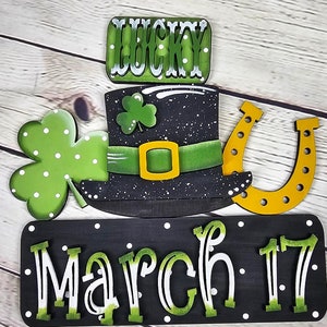 May include: A wooden sign with a black background and white polka dots. The sign says "March 17" in green and white letters. Above the sign is a black leprechaun hat with a green band and a gold buckle. There is a green shamrock on the hat and a gold horseshoe to the right. A green shamrock with white polka dots is to the left of the hat. A wooden sign with the word "LUCKY" in green and white letters is above the hat.