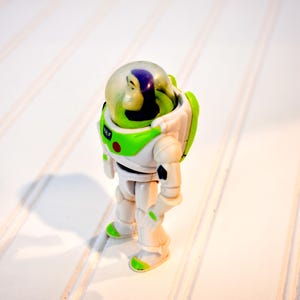 Może przedstawiać: Figurka akcji Buzz Lightyear, w białym i zielonym kombinezonie kosmicznym z przezroczystym hełmem. Zabawka ma ruchome kończyny i czerwony guzik na piersi. Figurka stoi na białej powierzchni.