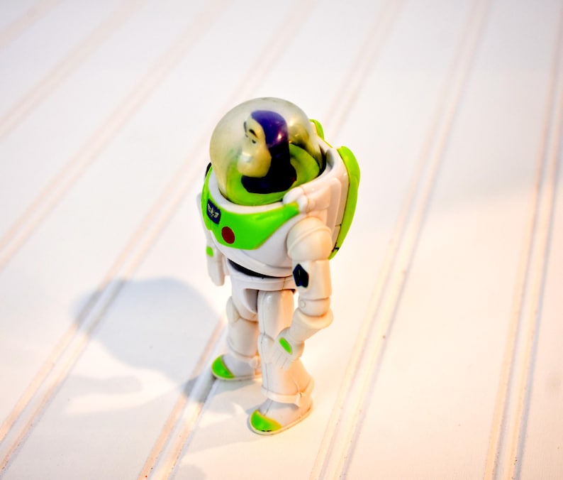 Może przedstawiać: Figurka akcji Buzz Lightyear, w białym i zielonym kombinezonie kosmicznym z przezroczystym hełmem. Zabawka ma ruchome kończyny i czerwony guzik na piersi. Figurka stoi na białej powierzchni.