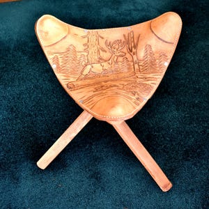May include: A light brown, three-legged folding stool with a leather seat. The seat features a carved forest scene with a deer. The legs are made of light-colored wood, and the stool is set against a dark green background.