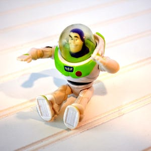 Może przedstawiać: Figurka Buzz'a Lightyear'a w pozycji siedzącej. Figurka ma biało-zielony skafander kosmiczny z przezroczystym hełmem. Ręce są wyciągnięte, a na piersi znajduje się czerwony guzik. Zabawka znajduje się na białej powierzchni.