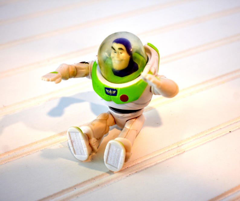 Może przedstawiać: Figurka Buzz'a Lightyear'a w pozycji siedzącej. Figurka ma biało-zielony skafander kosmiczny z przezroczystym hełmem. Ręce są wyciągnięte, a na piersi znajduje się czerwony guzik. Zabawka znajduje się na białej powierzchni.
