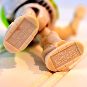 Może przedstawiać: Zbliżenie st&oacute;p figurki Buzz Lightyear. Podeszwy st&oacute;p są w kolorze beżowym z nadrukiem "MFG. FOR BURGER KING CORPORATION" i "&copy;PIXAR MADE IN CHINA AF-06-41". Figurka leży na białej powierzchni.